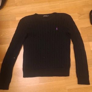 Svart stickad tröja från Ralph Lauren, Passar 152-158. - Snygg svart stickad tröja från Ralph Lauren med lila broderad logga på bröstet. Tröjan har en klassisk design med ribbade detaljer och långa ärmar. Perfekt för en stilren look. Funkar bäst nu på vintern eftersom den är varm och stickad. Pris kan diskuteras vid snabb affär.
