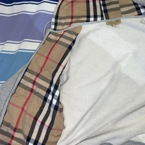 Grå huvtröja från Burberry - Säljer en stilren grå huvtröja från Burberry med klassiskt rutigt mönster på insidan av huvan. Tröjan har dragkedja och justerbara snören vid huvan. Perfekt för en avslappnad look.