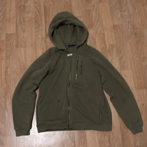 Olivgrön sailracing  - Säljer en olivgrön fleecejacka med huva och dragkedja framtill. Jackan har långa ärmar och en enkel design, perfekt för kyligare dagar. Den har en liten broderad detalj på bröstet.