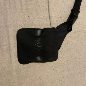 Svart axelväska från Hugo Boss - Snygg svart axelväska från Hugo Boss med justerbar rem och stilren design. Perfekt för att bära dina nödvändigheter med stil. Väskan har en  logotyp på framsidan.