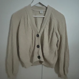 Beige stickad kofta från Lindex - Säljer en mysig beige stickad kofta från Lindex. Koftan har stora knappar framtill och långa ärmar med ribbade muddar. Perfekt för kyliga dagar!