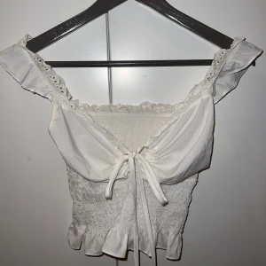 Vit blus med spetsdetaljer - Charmig vit blus med spetsdetaljer och volang. Den har en knytning framtill och är offshoulder med korta ärmar. Perfekt för en somrig look!