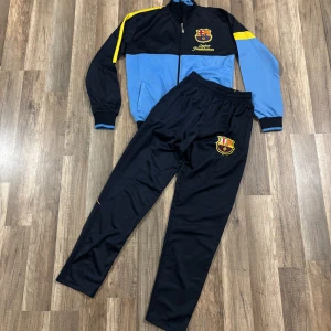 Blå och svart träningsjacka med barcelona märke - Snygg träningsjacka i blått och svart med klubbmärke på bröstet och texten 'Barça' på ryggen. Jackan har dragkedja och långa ärmar med gula detaljer. Perfekt för fotbollsfans!