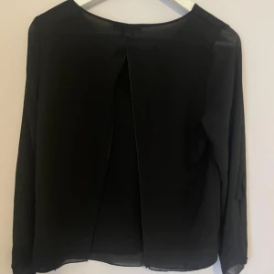 Svart blus från Italia Moda - Elegant och stilren svart blus från Italia Moda med långärmad design. Blusen har en unik omlott-detalj på ryggen.