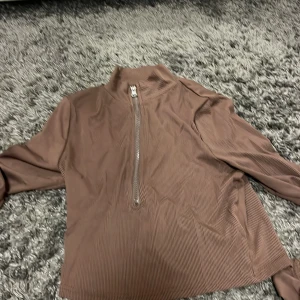 Brun tröja med dragkedja - Säljer en brun tröja med halv dragkedja och hög krage. Tröjan har långa ärmar och ett diskret randigt mönster. Perfekt för en avslappnad stil.