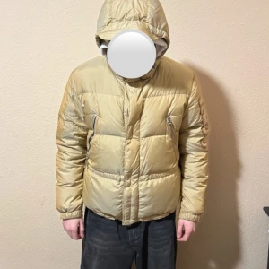 Beige dunjacka från Kejo  - Snygg beige japansk dunjacka från Kejo med broderade detaljer på ryggen och ärmen. Jackan har en huva och dragkedja framtill, perfekt för kyliga dagar. Den är varm och skön helt enkelt perfekt för vintern, jag har använt den som (tjej 170cm) lite oversized men passar lika bra som herr modell eller dam modell - svin snygg jacka som är svår att få tag på