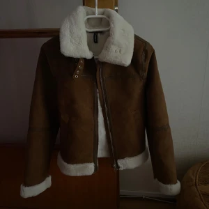 Brun jacka med foder från H&M - Säljer en snygg brun jacka från H&M med vit fuskpälsfoder. Jackan har en dragkedja framtill och en hög krage för extra värme. Perfekt för kyliga dagar!