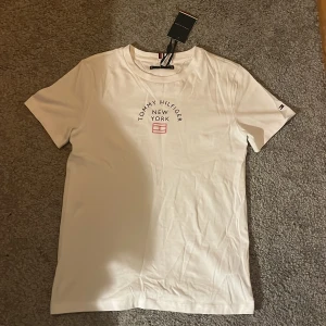 Vit t-shirt från Tommy Hilfiger - Säljer en stilren vit t-shirt från Tommy Hilfiger med tryck 'New York' på framsidan. T-shirten är kortärmad och har en klassisk passform. Perfekt för en avslappnad look. Alldrig använd. 