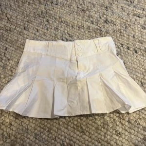 Vit kortkjol med knappar - Snygg vit kortkjol med veckad design och knappar framtill. Perfekt för en stilren look.