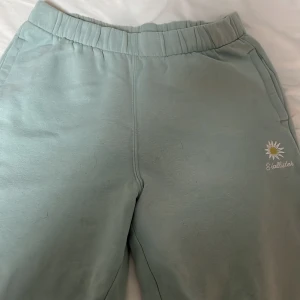 Gröna mjukisbyxor från Hollister - Säljer ett par gröna mjukisbyxor från Hollister med broderad sol och text på sidan. Byxorna har en ultra hög midja och elastiska muddar vid bensluten för extra komfort. Perfekta för en avslappnad stil.