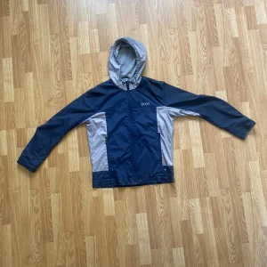 Blå och grå vindjacka från Hugo Boss - Snygg vindjacka från Hugo Boss i blått och grått med huva och dragkedja. Jackan har en vattenavvisande yta och är perfekt för blåsiga dagar. Den har långa ärmar och en stilren design med logotyp på bröstet och ryggen.