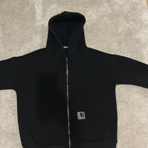 Svart hoodie från Carhartt - Säljer en svart hoodie från Carhartt med dragkedja och fickor framtill. Perfekt för en avslappnad stil. Tillverkad i bomull för extra komfort.