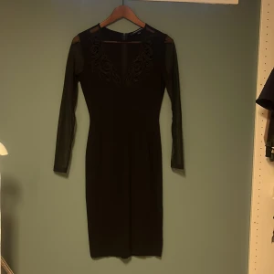 Svart klänning från French Connection - Elegant svart klänning från French Connection med långa ärmar i transparent mesh och prickigt mönster. Klänningen har en vacker spetsdetalj vid halsringningen och en dragkedja i ryggen. Perfekt för en stilren look.
