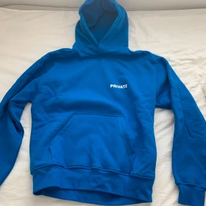 Blå hoodie med texten 'PRIVATE' - Säljer en blå hoodie med texten 'PRIVATE' på bröstet. Den har en klassisk känguruficka och en bekväm huva. Perfekt för en avslappnad stil.