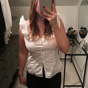 Vit blus med spetsdetaljer - Säljer en söt vit blus med spetsdetaljer och knappar framtill. Blusen har en v-ringad design och är ärmlös, perfekt för varmare dagar. Passar bra till både jeans och kjol.