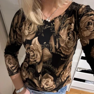 Svart topp med rosor - Snygg svart topp med stora bruna rosor och glittriga detaljer. Toppen har trekvartsärm och en normal passform. Perfekt för att ge en elegant touch till din outfit.