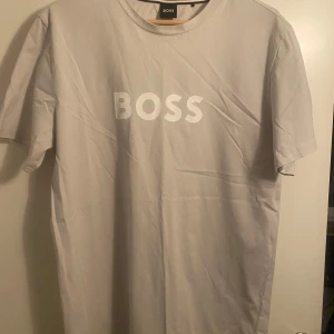 Grå t-shirt från BOSS - Snygg gråbeige  t-shirt från BOSS i ekologisk bomull med solskydd. Regular fit med korta ärmar och stort BOSS-tryck på framsidan. Perfekt för en stilren och bekväm look.