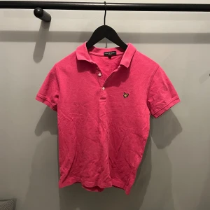 Rosa pikétröja från Lyle & Scott - Snygg rosa pikétröja från Lyle & Scott med klassisk krage och knappar. Perfekt för en avslappnad stil med en liten broderad logga på bröstet.