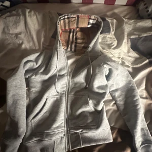Grå hoodie från Burberry - Säljer en stilren grå hoodie från Burberry med klassiskt rutigt mönster i luvan. Tröjan har dragkedja och fickor framtill, perfekt för en avslappnad look. Passar bra till både vardag och chill.
