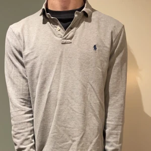 Grå långärmad pikétröja från Ralph Lauren - Säljer en stilren grå långärmad pikétröja från Ralph Lauren med klassisk krage och knappdetalj. Den har en broderad logotyp på bröstet i blått. Perfekt för en avslappnad men ändå snygg look.
