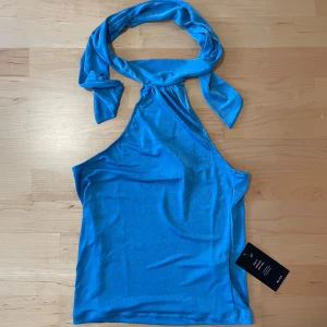 Blå oneshoulder topp oanvänd M - Snygg blå oneshoulder topp från NA-KD med knytband runt halsen. Perfekt för en somrig look. Tillverkad i mjukt material som ger en bekväm passform.