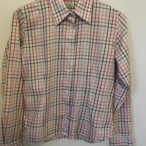 Rutig skjorta från Sand - Snygg rutig skjorta från Sand i flera färger som rosa, blå och svart. Skjortan har långa ärmar och knappar framtill. Perfekt för en stilren look. Bredd skuldra -Skuldra 39 cm Bröst 46 cm Längd från axel 56 cm Längd från armhåla 35 cm