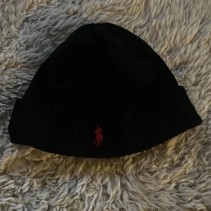 Svart mössa med röd logga - Snygg svart mössa från Polo Ralph Lauren med en röd broderad logga framtill. Perfekt för kyliga dagar. Bra skick