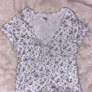 Blommig kortärmad topp  - Söt blommig topp från PAC-SUN i vitt med små blommor i lila och blå. Toppen är öppen fram med knytning. Aldrig använd.