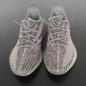 Adidas Yeezy Boost 350 Beluga 2.0 - Säljer ett par Adidas Yeezy Boost 350 V2 i grått med mönstrad ovandel och texten 'SPLY-350' i orange. Skorna har en ribbad sula och snörning för en bekväm passform.