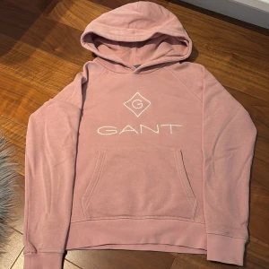 Rosa hoodie från GANT - Säljer en snygg rosa hoodie från GANT med vit logotyp på bröstet. Tröjan har en klassisk känguruficka och en bekväm huva. Perfekt för en avslappnad stil.