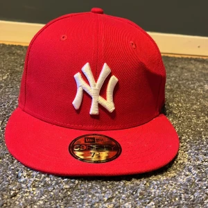 Röd keps från New Era - Snygg röd keps från New Era med broderad vit logga framtill. Perfekt för att ge din outfit en sportig touch. Tillverkad i polyester och har en klassisk passform.Den har en väldigt liten buckla på sidan som knappt syns.Därför säljs den för mindre.