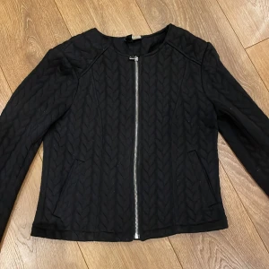 Svart quiltad jacka från H&M Divided - Snygg svart quiltad jacka från H&M Divided med dragkedja framtill. Jackan har en stilren design med långärmade ärmar och ett diskret mönster. Perfekt för en trendig look.