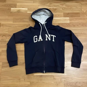 Mörkblå hoodie från GANT - Säljer en snygg mörkblå hoodie från GANT med dragkedja och vit text på bröstet. Den har en justerbar huva med vita snören och långa ärmar. Aningens urtvettad. Perfekt för en avslappnad stil.