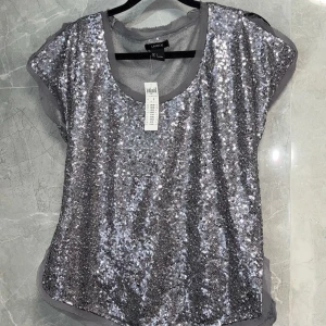 Glittrig silver topp från Lindex  - Snygg glittrig topp i silver från Lindex . Perfekt för festliga tillfällen med korta ärmar och rund halsringning. Toppen är täckt av paljetter som ger en extra glamorös känsla.