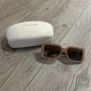 Beige/rosa solglasögon från Michael Kors - Snygga solglasögon från Michael Kors i en trendig beige/ rosa färg. De har en fyrkantig form och logotypen tryckt på bågarna. Perfekta för att ge en stilren touch till din look. Kommer med ett vitt fodral för säker förvaring.