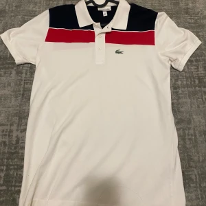 Vit pikétröja från Lacoste - Snygg vit pikétröja från Lacoste med röd och marinblå rand över bröstet. Tröjan har korta ärmar och en klassisk krage med knappar. Lacoste-loggan är broderad på framsidan och märket är tryckt på baksidan.