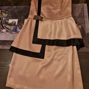 Champagnefärgad klänning med svarta detaljer - Elegant champagnefärgad klänning med svarta kanter och en dekorativ rosett i midjan. Klänningen har en asymmetrisk design och är axelbandslös, vilket ger en stilren look.storlek 38