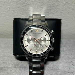 Silverfärgad klocka från Emporio Armani - Stilren klocka från Emporio Armani med silverfärgad urtavla och armband. Klockan har en tachymeter och flera små urtavlor för extra funktionalitet. Perfekt för den som vill ha en elegant och funktionell accessoar.