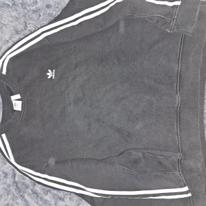 Svart sweatshirt från Adidas - Säljer en klassisk svart sweatshirt från Adidas med de ikoniska tre ränderna längs ärmarna. Tröjan har en rund halsringning och det vita Adidas-logotypen på bröstet. Perfekt för en sportig och avslappnad stil.