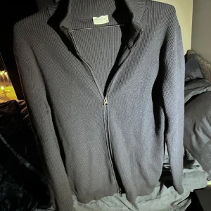 Mörkblå ribbad kofta från John Henric - Säljer en stilren mörkblå ribbad kofta från John Henric. Koftan har en dragkedja framtill och långa ärmar, perfekt för kyligare dagar. Passar både till vardags och mer uppklädda tillfällen. Köpt för 1000