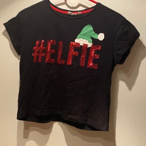 Svart t-shirt med julmotiv från KappAhl - Säljer en svart t-shirt från KappAhl med texten '#ELFIE' i röda paljetter och en grön tomteluva. Perfekt för julens festligheter! Kortärmad och bekväm passform. Super fin i st 158/164 passar XS. 