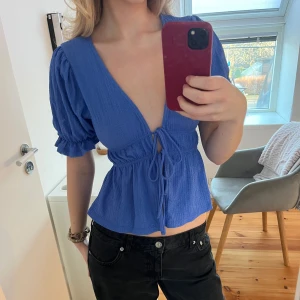 Blå v-ringad blus med knyt - Snygg blå blus med djup v-ringning och knyt framtill. Blusen har korta puffärmar och en lätt volang vid nederkanten. Perfekt för en stilren look.