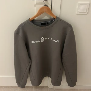 Grå sweatshirt från Sail Racing - Snygg grå sweatshirt från Sail Racing med vit logotyp på bröstet och en mindre logotyp på ryggen. Tröjan har en rund halsringning och långa ärmar, perfekt för en avslappnad stil.