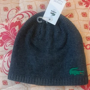 Grå ribbad mössa från Lacoste - Snygg grå ribbad mössa från Lacoste med det ikoniska gröna krokodilemblem broderat på sidan. Perfekt för kyliga dagar och en stilren look.