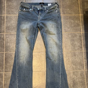 Blå jeans från True Religion - Säljer ett par snygga blå jeans från True Religion, modell Joey Low Rise Flare. De har en låg midja och utsvängda ben. Jeansen har klassiska fem fickor och dekorativa sömmar på bakfickorna.