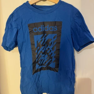 Blå t-shirt från Adidas 152 - Säljer en blå t-shirt från Adidas med ett stort tryck på framsidan. T-shirten är kortärmad och har en rund halsringning. Perfekt för sportiga aktiviteter eller en avslappnad stil.