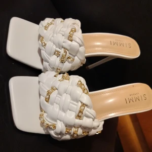 Vita klackskor från Simmi London - Stilrena vita klackskor från Simmi London med flätad design och gulddetaljer. Skorna har en fyrkantig tå och en elegant klack, perfekt för att lyfta din stil. 