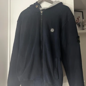 Mörkblå hoodie från Stone Island - Säljer en snygg mörkblå hoodie från Stone Island med dragkedja och huva. Jackan har märkets ikoniska logga på ärmen och en liten patch på bröstet. Perfekt för en casual look. På tröjan står det att den är i XL men jag skulle mer säga att den är i M. Pris går att diskutera