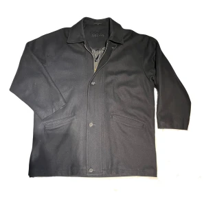 Marinblå ull kappa  - Snygg och stilren ull kappa i marinblå färg från Rappson. Kappan har en rak passform med fickor framtill och en dragkedja samt knappar i fram. Kragen är uppfällbar. Detaljerna är diskreta och ger kappan ett elegant utseende. Har även två fickor på insidan. Står ingen storlek i kappan men skulle säga någonstans mellan XL och XXL