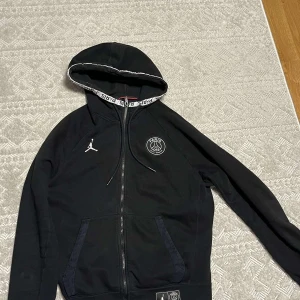  Jordan x PSG Ziphoodie - Säljer en svart hoodie från Jordan x PSG med dragkedja och broderad logga på bröstet. Hoodien har långa ärmar och en justerbar huva med snören. Perfekt för dig som gillar sportig stil och vill ha något bekvämt och snyggt.